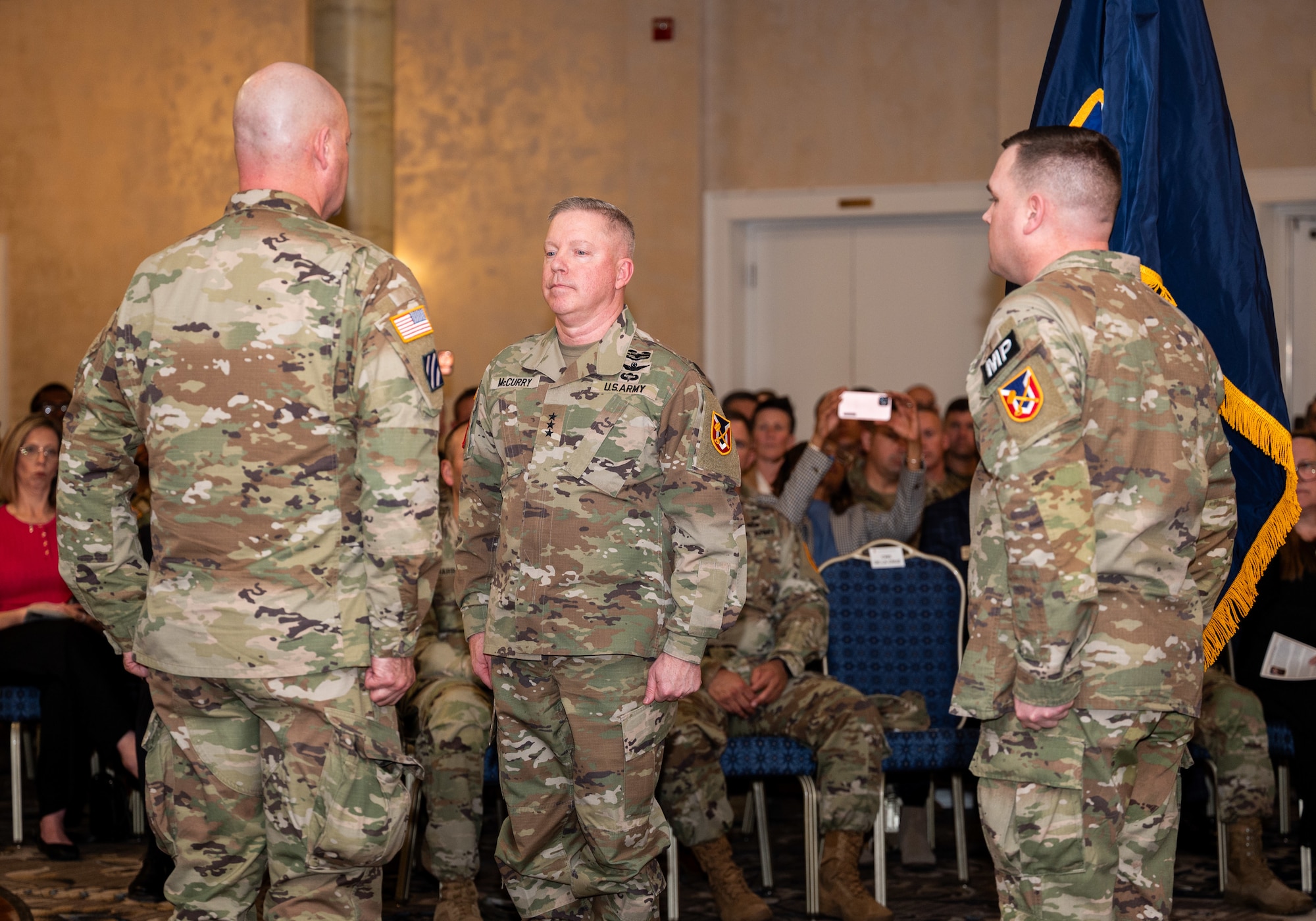 Three men in soldier uniforms stand at the position of attention in a ceremony room