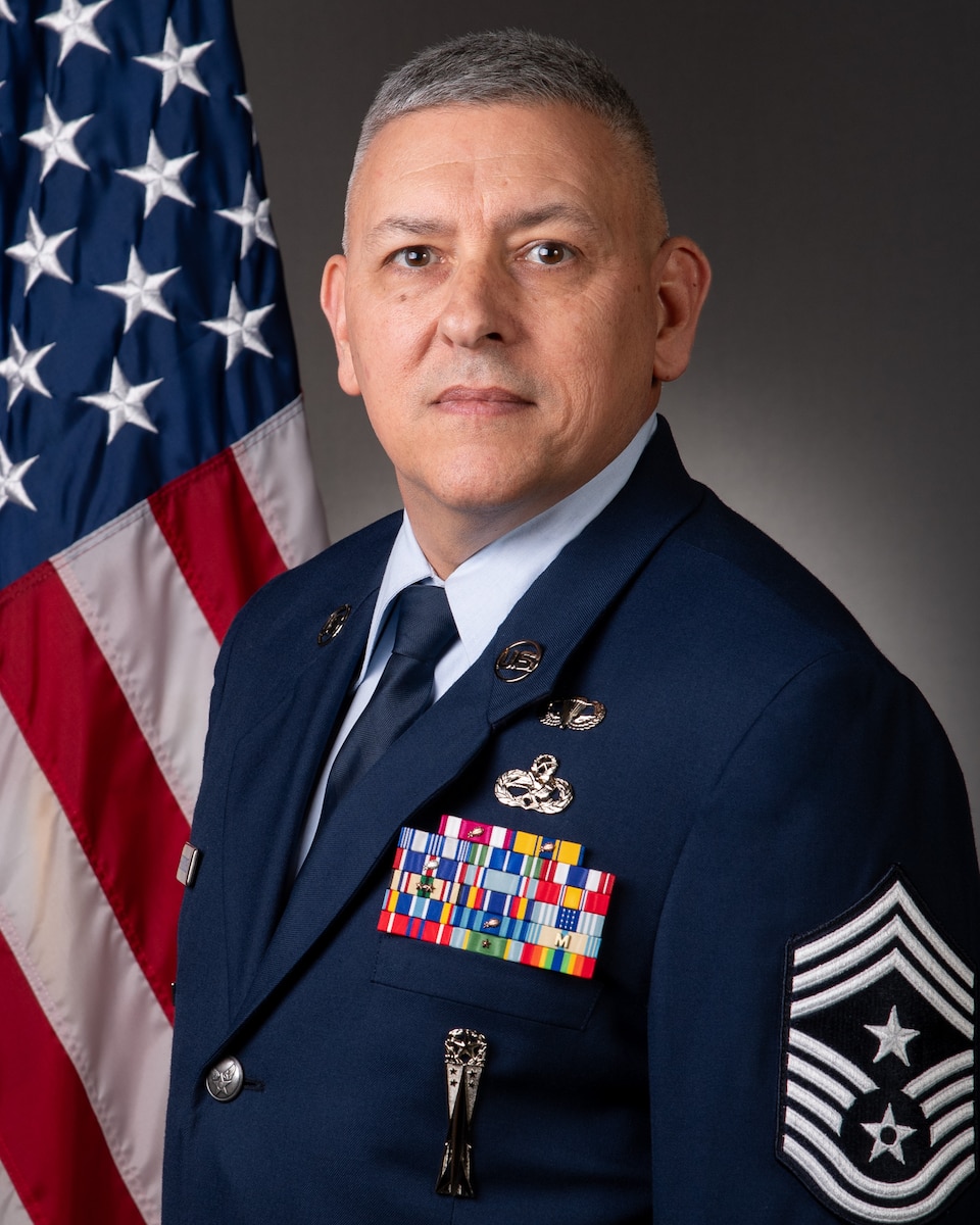 Official portrait of Chief Master Sgt. Jasen E. Hendricksen in service dress with U.S. American flag draped behind right shoulder.