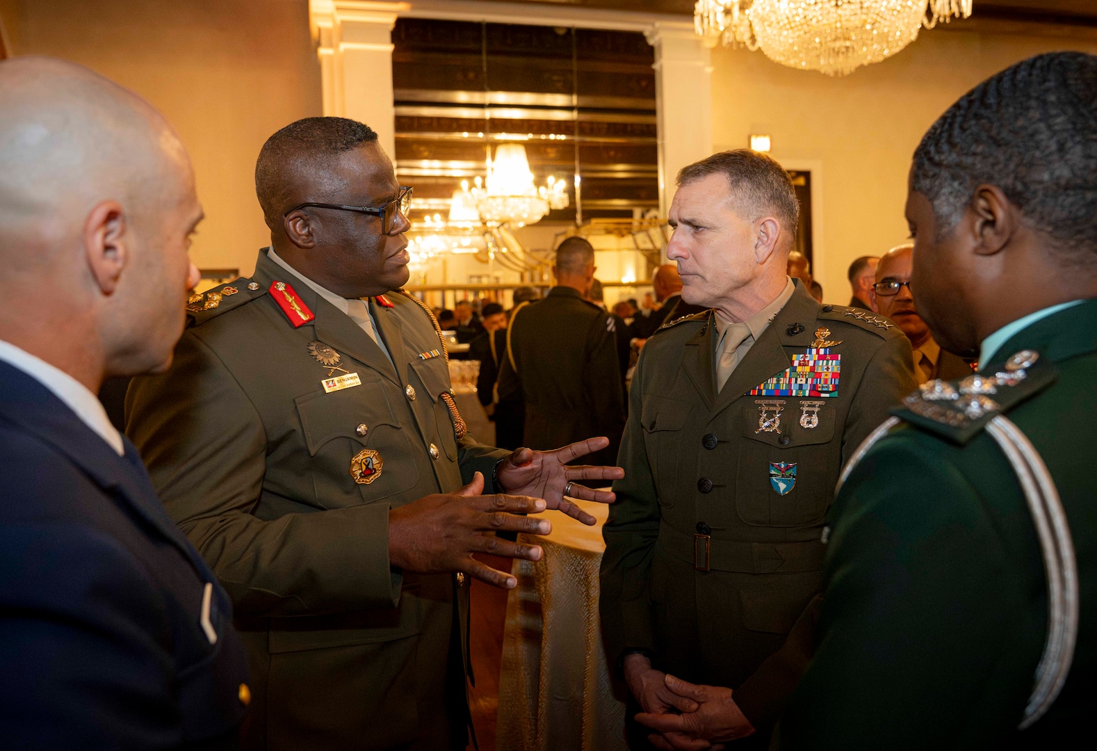A standing man in a brown military uniform speaks with a three other military personnel.