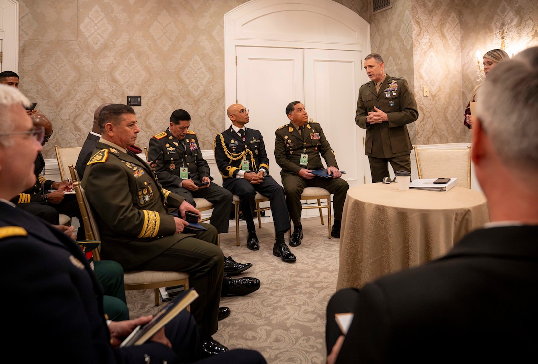 A standing man in a brown military uniform speaks with a group of sitting military personnel.
