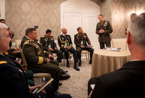 A standing man in a brown military uniform speaks with a group of sitting military personnel.