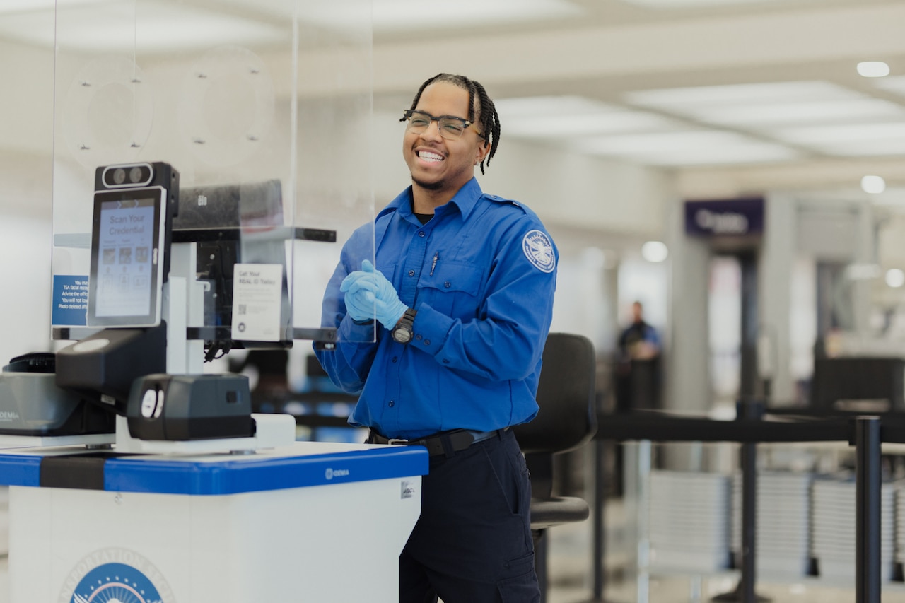 A smiling man in glasses wearing a long-sleeved blue shirt and black pants, stands behind a counter with a plexiglass shield hanging over it in a large terminal; electronic equipment is in the foreground.