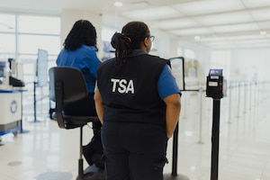 A pair of individuals, one sitting on an elevated chair to the left and wearing a blue shirt, and another standing to the right wearing a blue shirt and short-sleeved vest, are in a large terminal with their backs to the camera.