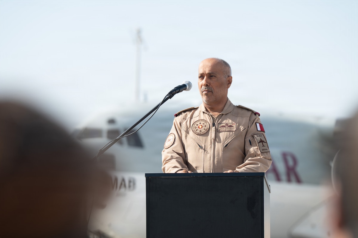 Qatar Emiri Air Force Staff Col. Pilot Mubarak Hasan Albin Ali speaks into a microphone from behind a podium. A Qatar Airways aircraft is behind him