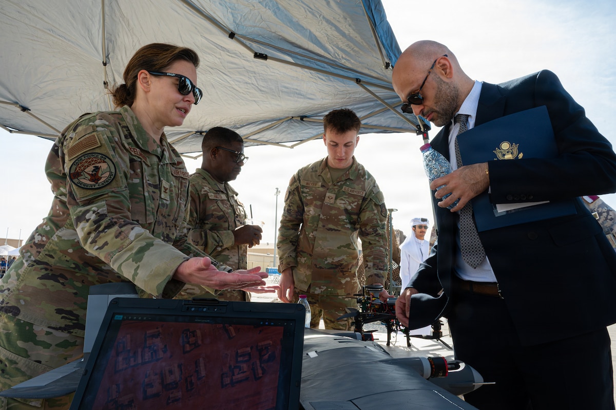 An Airman gestures to an unidentifiable object on a table under a canopy while Mo Barghouty looks on. Other Airmen are in the background showing someone a drone.