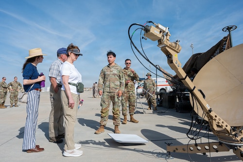 Three people in civilian clothes stand and look at a satellite dish while two Airmen talk to them