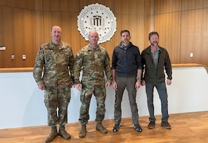 Two men in camouflage military uniforms and two men in business casual attire pose for a photo in a room. There is an emblem on the back wall that reads "Department on Justice, Federal Bureau of Investigations."