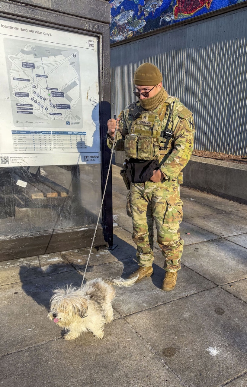 A man dressed in a camouflage military uniform and sunglasses holds the leash of a small fluffy white dog while standing next to a Metro station.