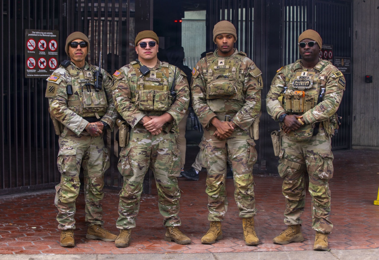 Four men in camouflage military uniform pose for a photo in front of a Metro station.