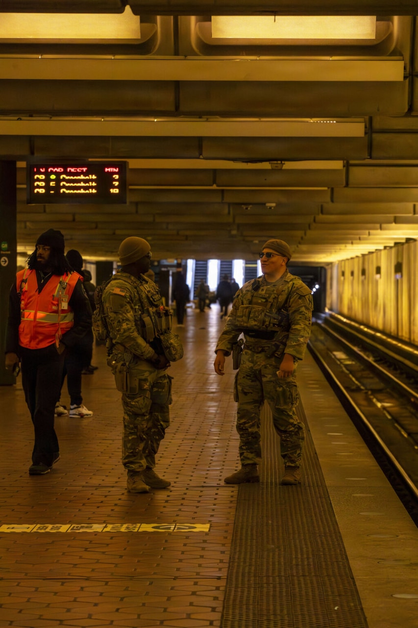 Two men in camouflage military uniform stand near the train tracks in an underground Metro station. Another man, wearing a Metro employee uniform and reflective vest, walks by.