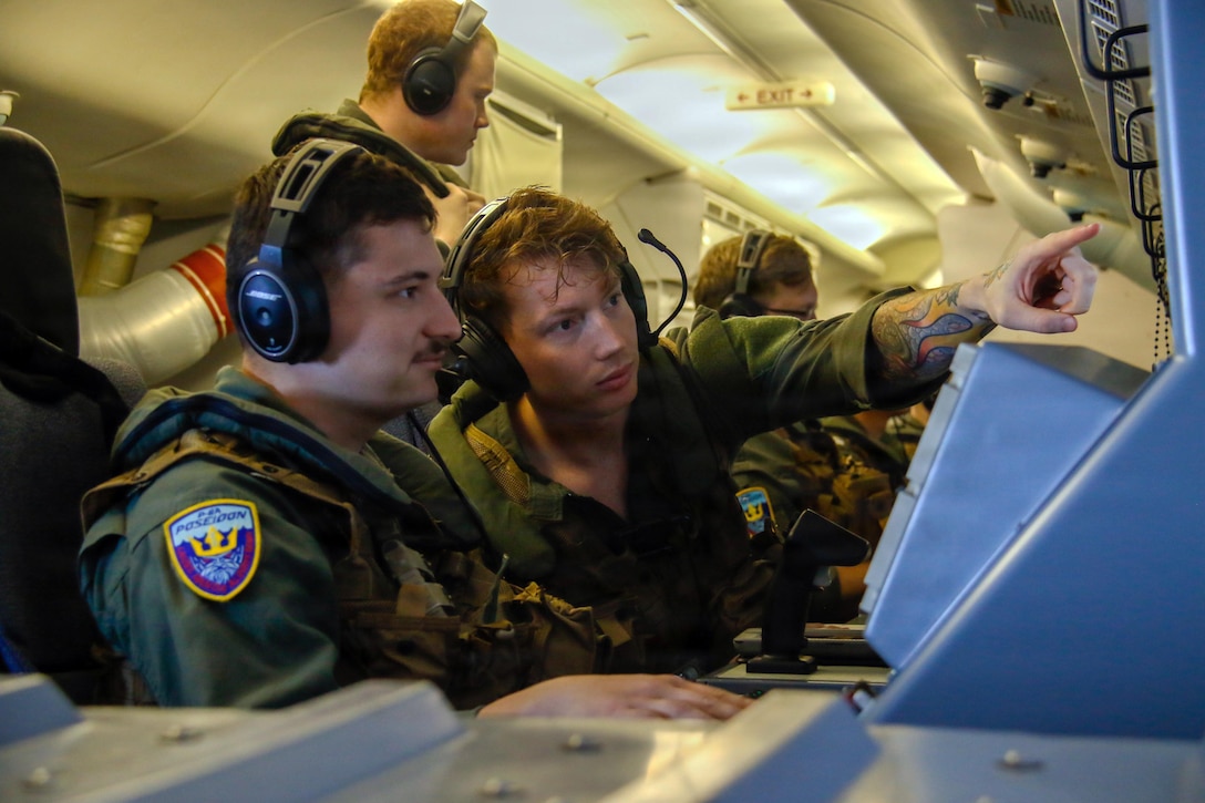 Two people wearing flight suits and headsets with a microphone attached sit and look at a screen; several people in similar attire are in the background.