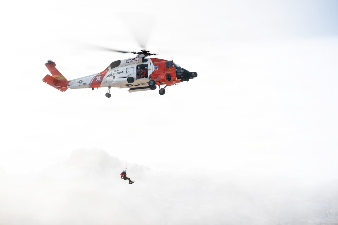 A Coast Guardsman is hoisted to an airborne helicopter in a bright, cloudy sky.