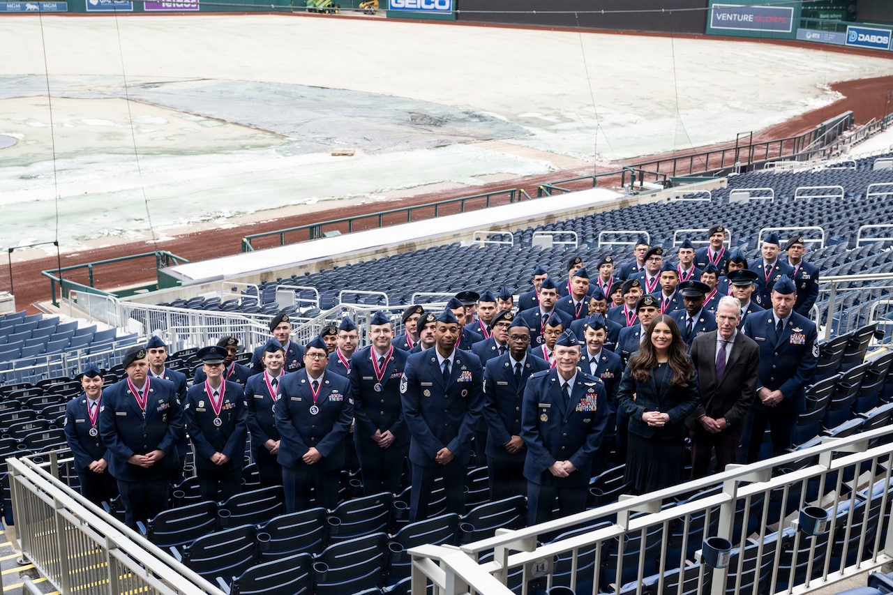 More than a dozen men and women wearing formal military uniforms stand in a stadium, posing for a picture around a man and a woman in business attire. A snow-covered baseball field is in the background.