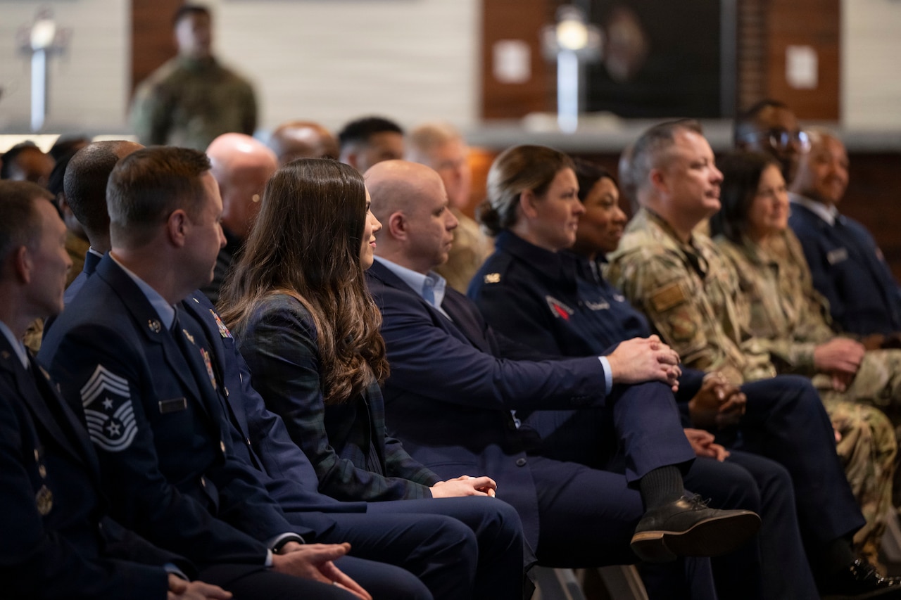 More than a dozen people, wearing a mix of business attire, formal military uniforms and camouflage military uniforms, sit in an indoor ceremony.