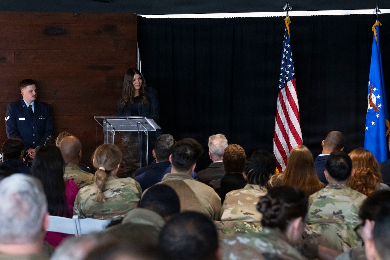 A woman in business attire stands behind a lectern and speaks to more than a dozen people wearing a mix of business attire and camouflage military uniforms. A man in a formal military uniform stands to her right, and on her left are an American flag and a blue flag with an eagle encircled by stars in the center.