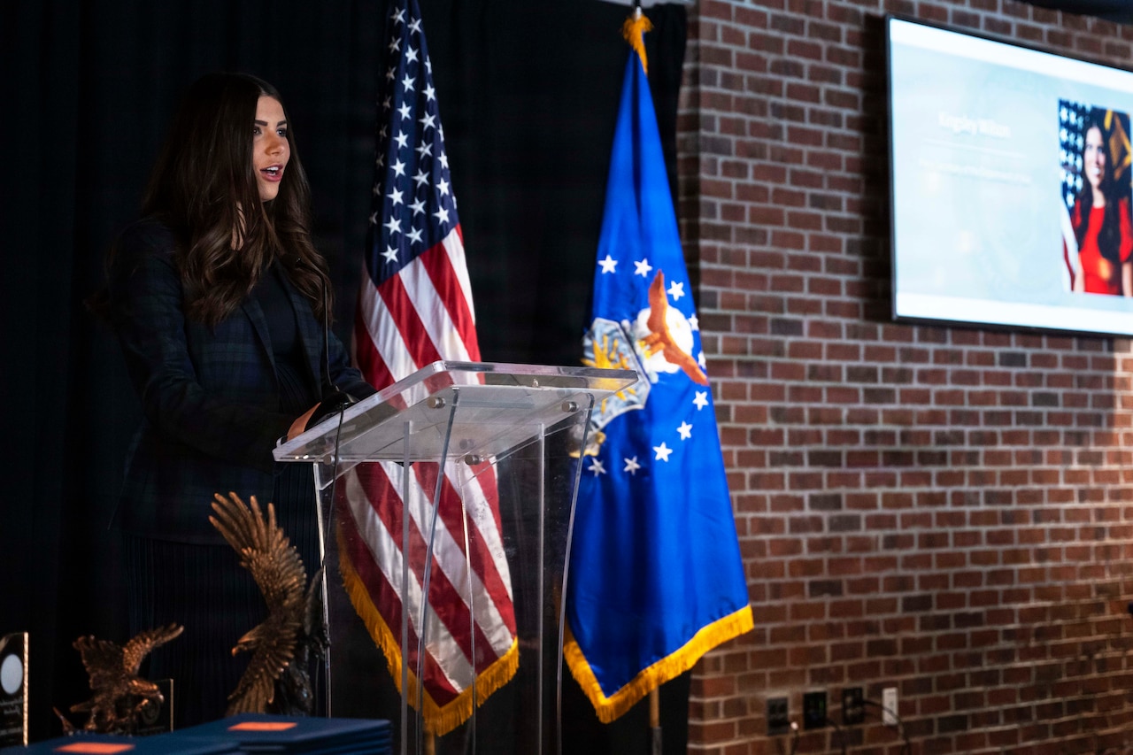 A woman in business attire stands behind a lectern and speaks. On her left are an American flag, a blue flag with an eagle encircled by stars in the center and a brick wall with a monitor hanging from it.