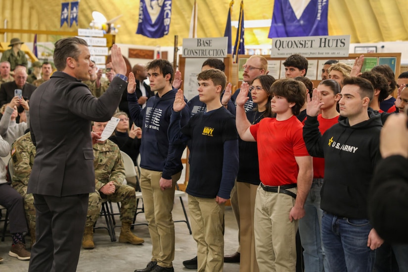 A man in a business suit, his right hand raised, stands in front of a group of approximately 15 young men and women in casual attire, also raising their right hands, indoors with a seated group observing.