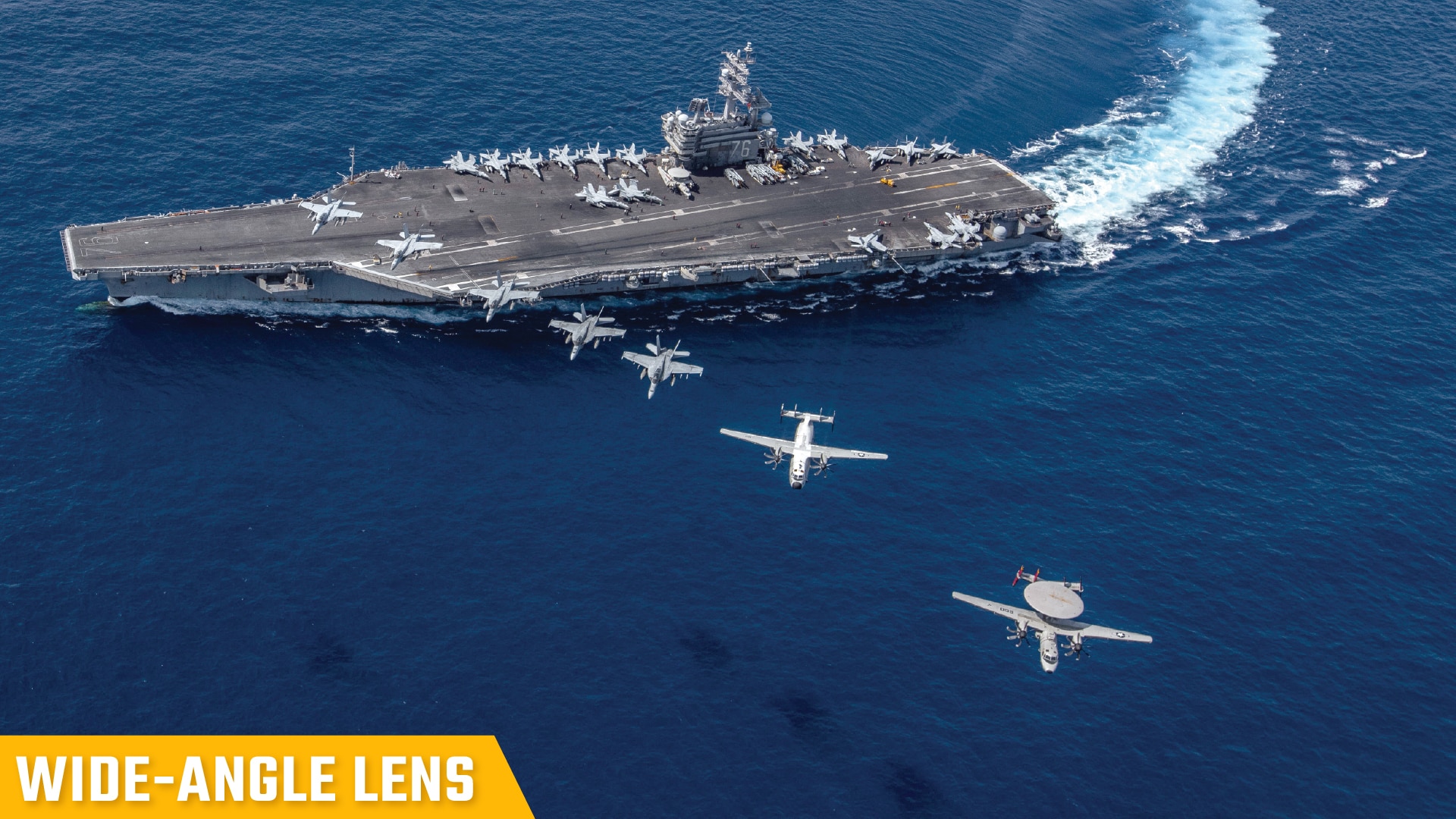 An aerial view captures a group of aircraft flying in a line over a sailing aircraft carrier, with the label "wide-angle lens" in the bottom left corner.