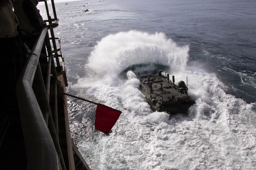 An amphibious combat vehicle splashes into the water after departing from a large ship on the left. There are several other combat vehicles floating in the background.