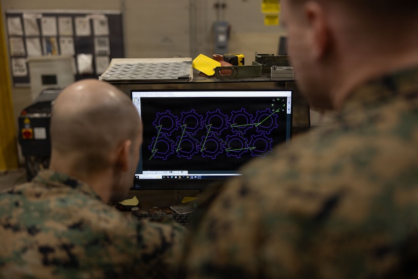 Two men in camouflage military uniforms look at a computer screen with a design for several metal washers displayed on it.
