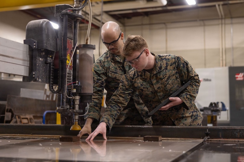 Two men in camouflage military uniforms and safety glasses put their hands on a piece of metal under a machine. The men are working in a large maintenance building.