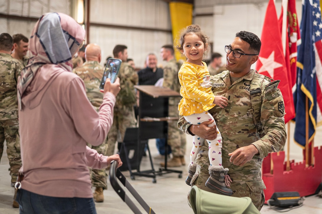 A loved takes a picture of a soldier holding a child as fellow soldiers gather to the left while flags sit to the right.