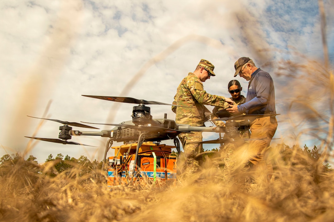 A drone sits in a grassy field as three people gather around a remote control on a sunny day.