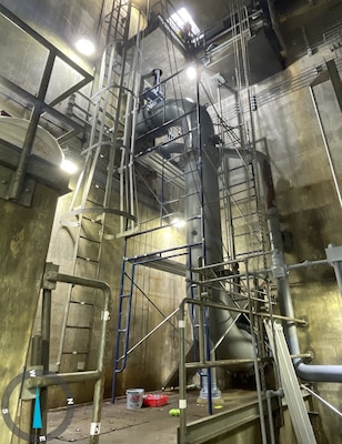 A large grey metal pipe with metal scaffolding surrounding it with concrete walls and floor in the background.