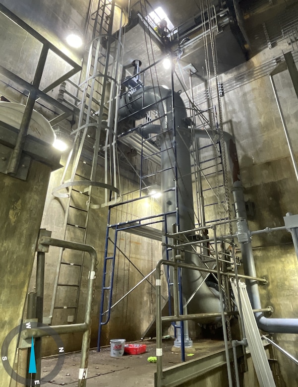 A large grey metal pipe with metal scaffolding surrounding it with concrete walls and floor in the background.