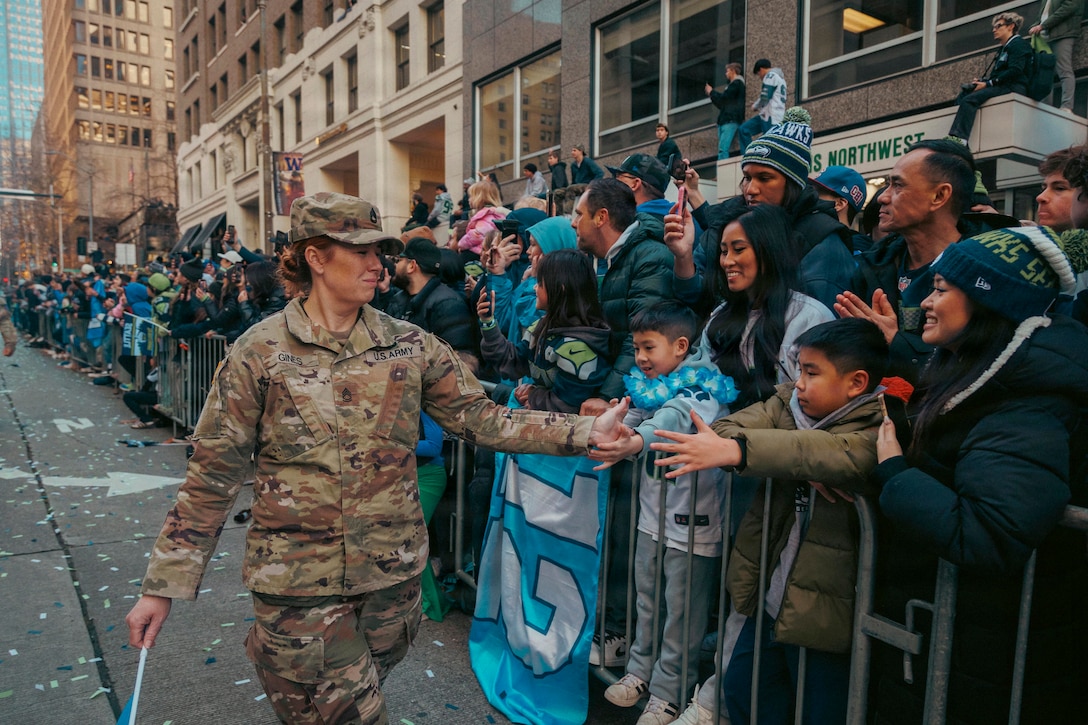 A walking soldier high-fives young fans standing in a large crowd behind a barrier near tall buildings during a parade.