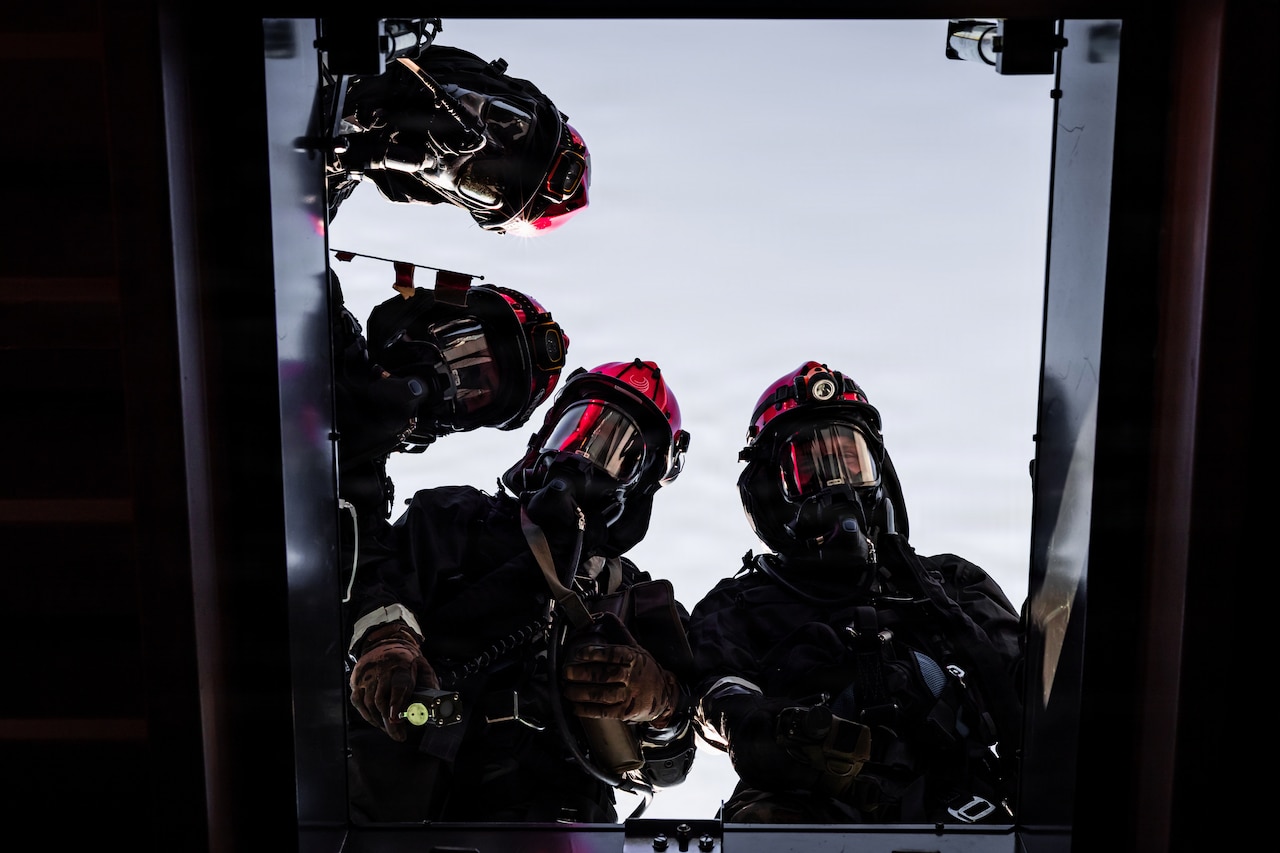 Four people dressed in firefighter gear look down into a dark hole.