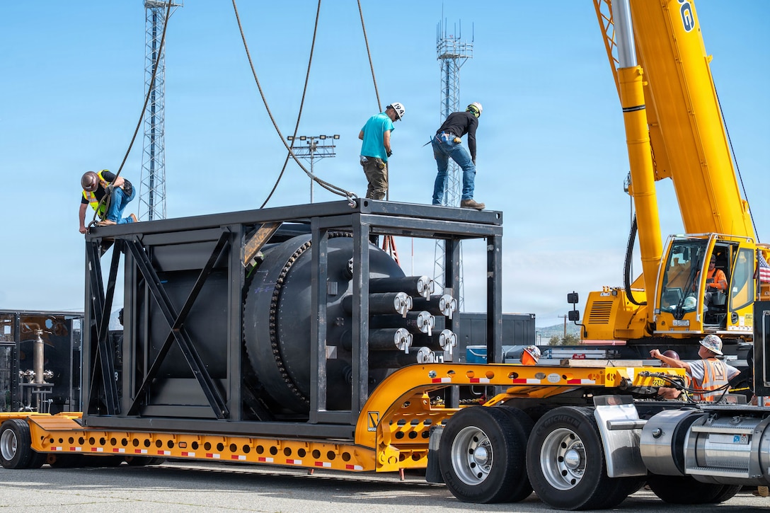 A containerized nuclear power reactor sits in a steel frame on a truck bed as three workers attach hooks onto it against a blue sky.