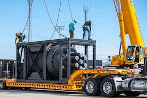 A containerized nuclear power reactor sits in a steel frame on a truck bed as three workers attach hooks onto it against a blue sky.