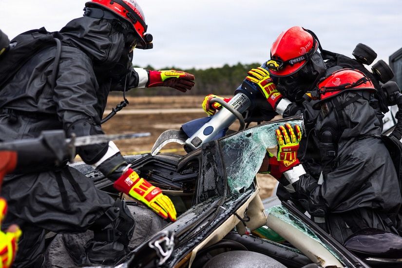 Three people dressed in firefighter gear attempt to pry open a wrecked car using a large metal device.