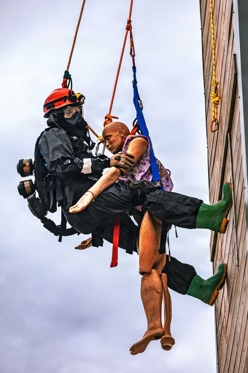 A person dressed in firefighter gear rappels down a wall with a mannequin.