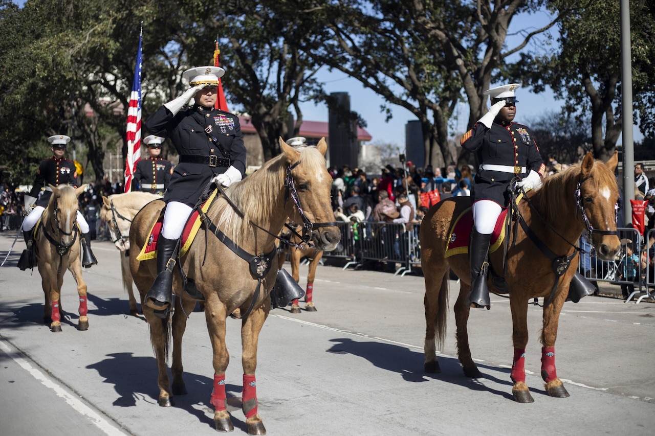Two men in military dress uniforms salute as they sit stoically on horses while on a parade route. Three other people in similar attire on horses are behind them carrying flags.