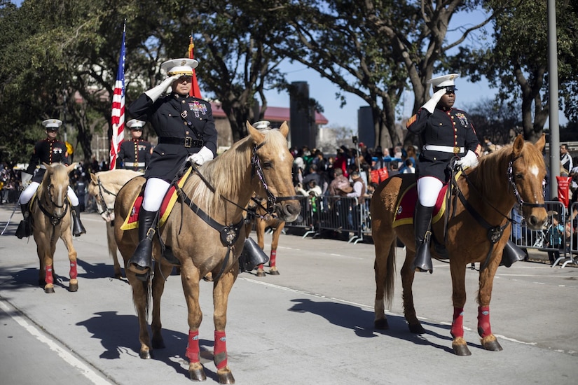 Two men in military dress uniforms salute as they sit stoically on horses while on a parade route. Three other people in similar attire on horses are behind them carrying flags.