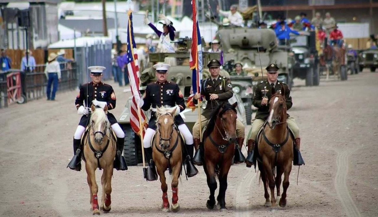 Four men in military dress uniforms on horseback march in unison along a dirt path. Military tanks topped with people waving from them move behind them in a line.