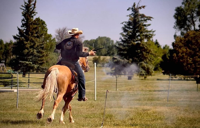 A man on horseback wearing a cowboy hat shoots a pistol from his left hand while riding through grass.