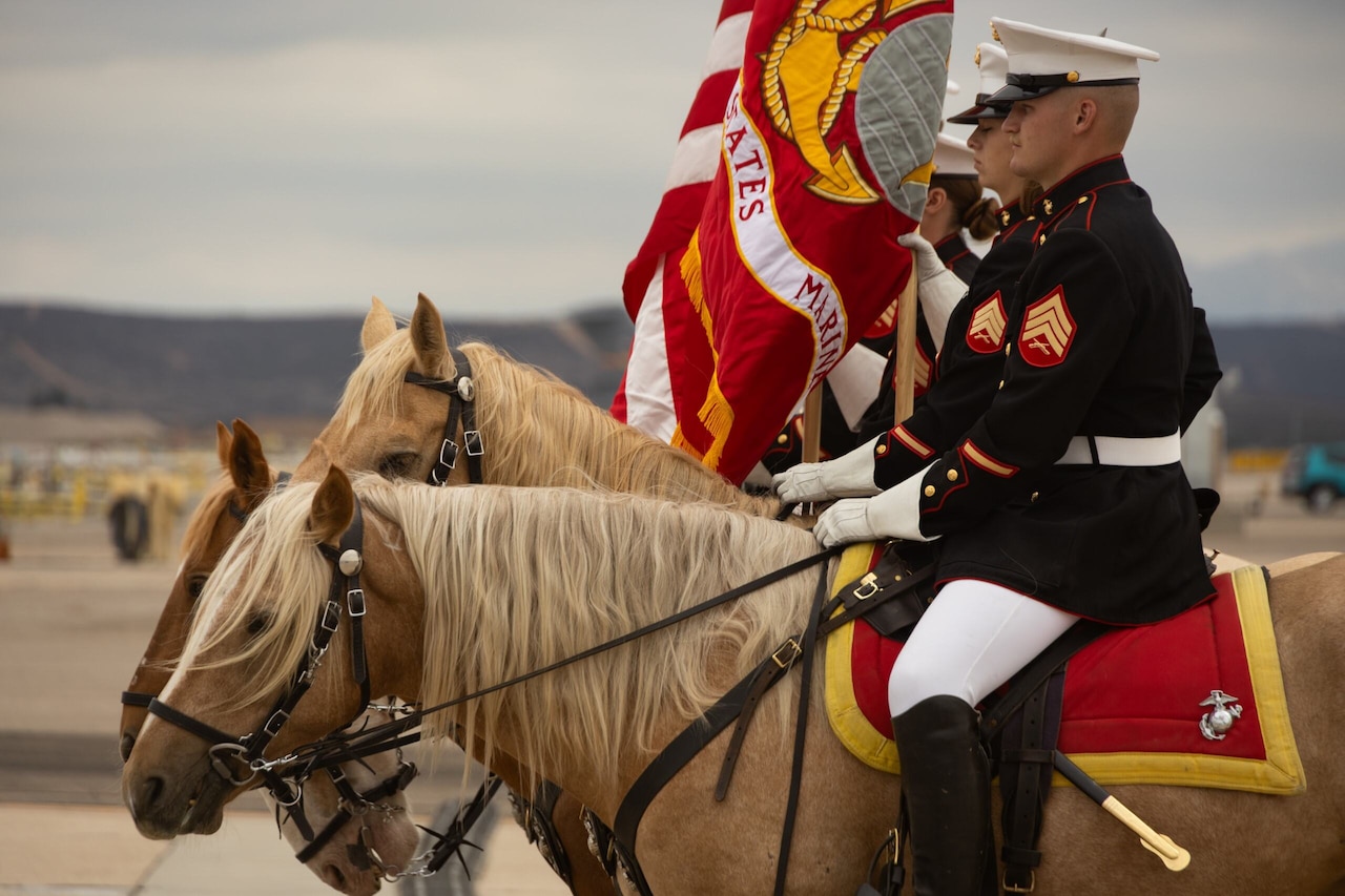 A side view of a man in a military dress uniform sitting on a horse. Three other riders in similar attire sit on horseback directly to his right. Two of them are carry flags on poles.