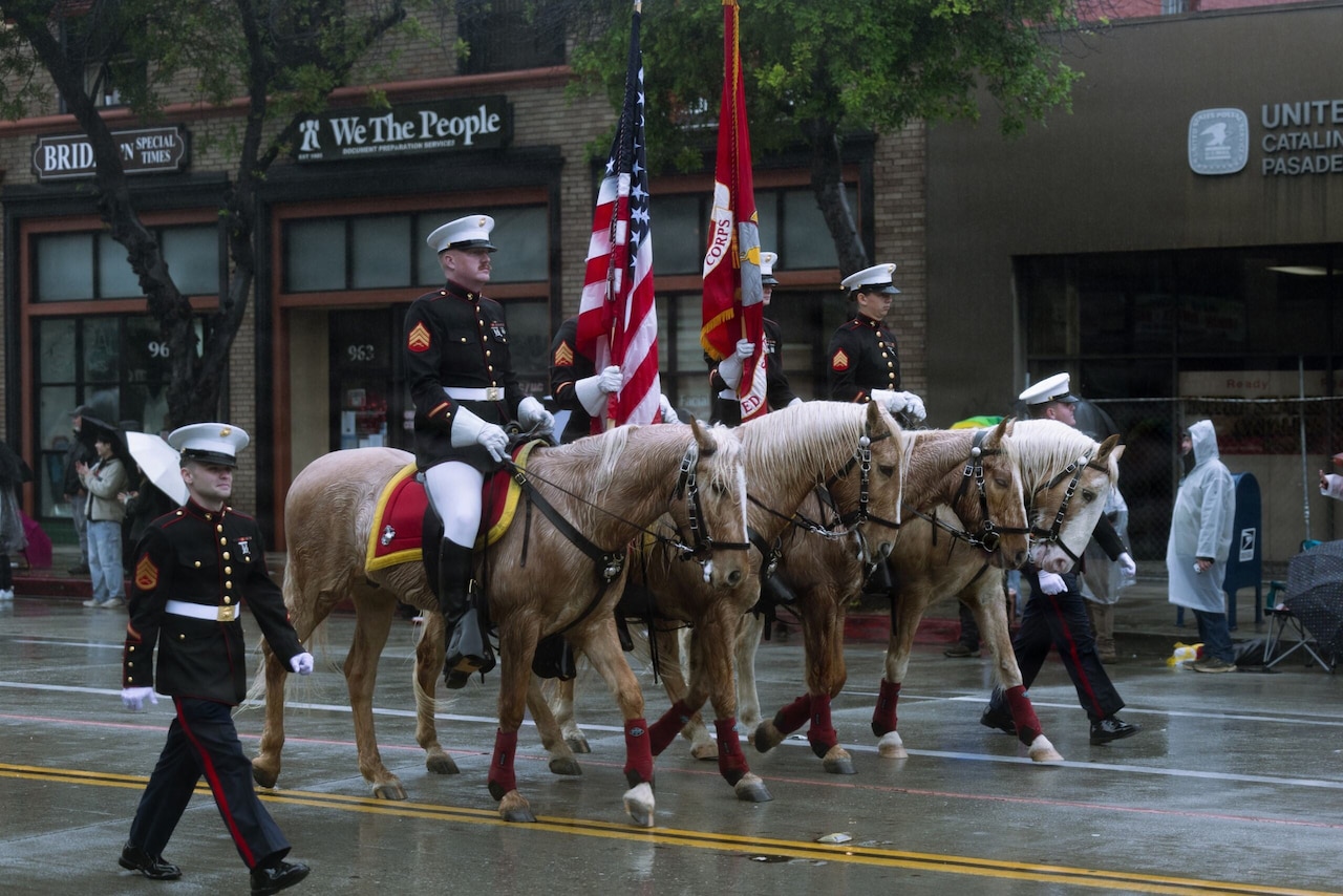 Four people in military dress uniforms on horseback move side by side on a paved road. Two people in similar attire walk beside the horses on the outside of the formation.