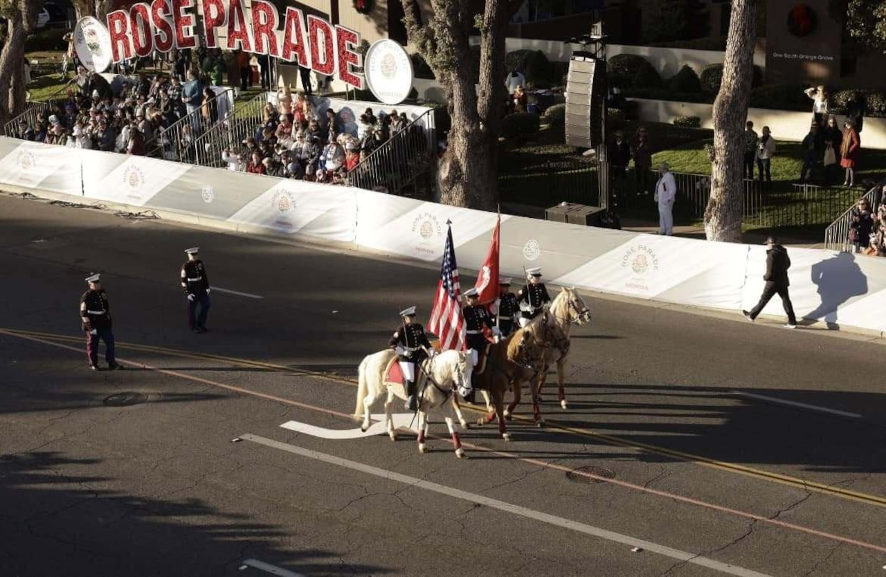 Four people in military dress uniforms on horseback march through an empty street. The two people in the middle hold flagpoles. Two people in similar attire walk in the distance behind them. In the background are parade stands with a large sign that reads “Rose Parade.”