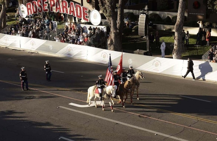 Four people in military dress uniforms on horseback march through an empty street. The two people in the middle hold flagpoles. Two people in similar attire walk in the distance behind them. In the background are parade stands with a large sign that reads “Rose Parade.”
