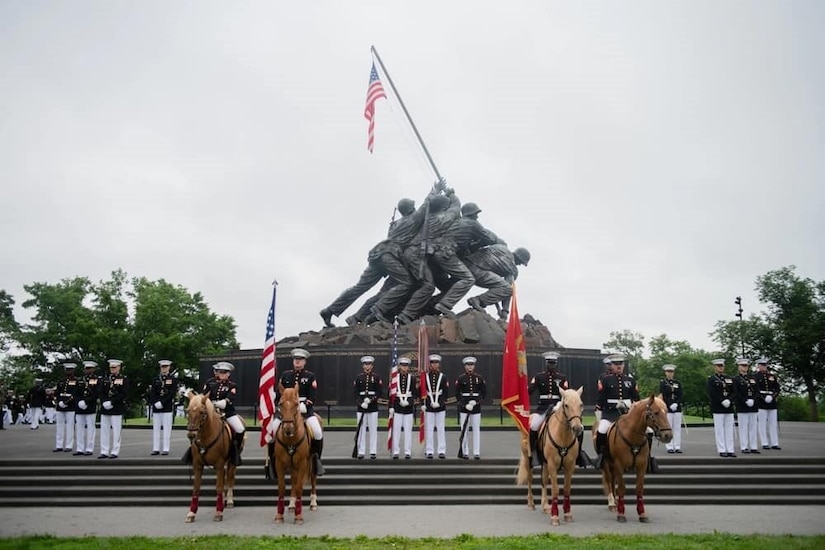 About two-dozen people wearing military dress uniforms pose for a photo in front of a large monument of men putting up a U.S. flag. Four riders in similar attire on horseback stand in front of the Marines.