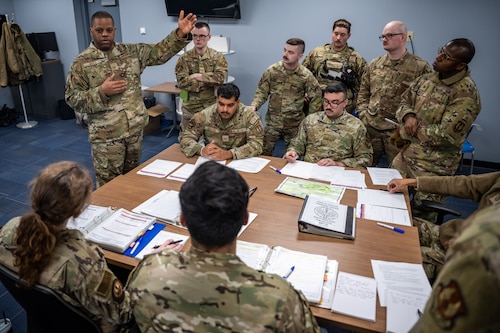 A group of men and women, wearing camouflage military uniforms, sit around a table covered in binders, maps, and documents.