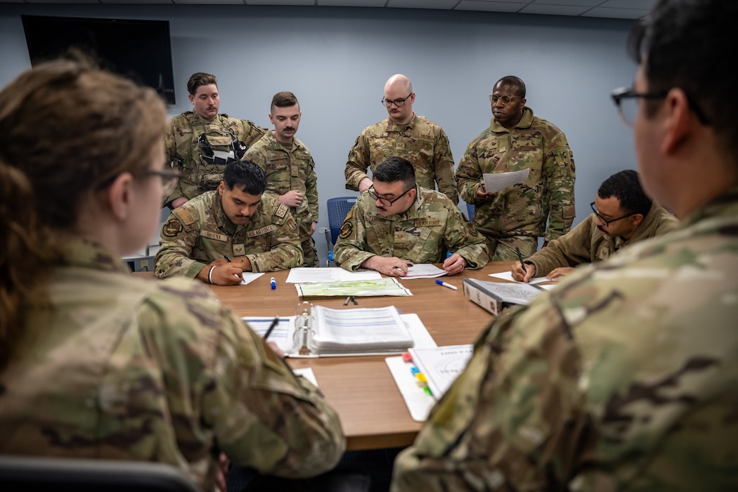 A group of men and women, wearing camouflage military uniforms, sit around a table covered in binders, maps, and documents.