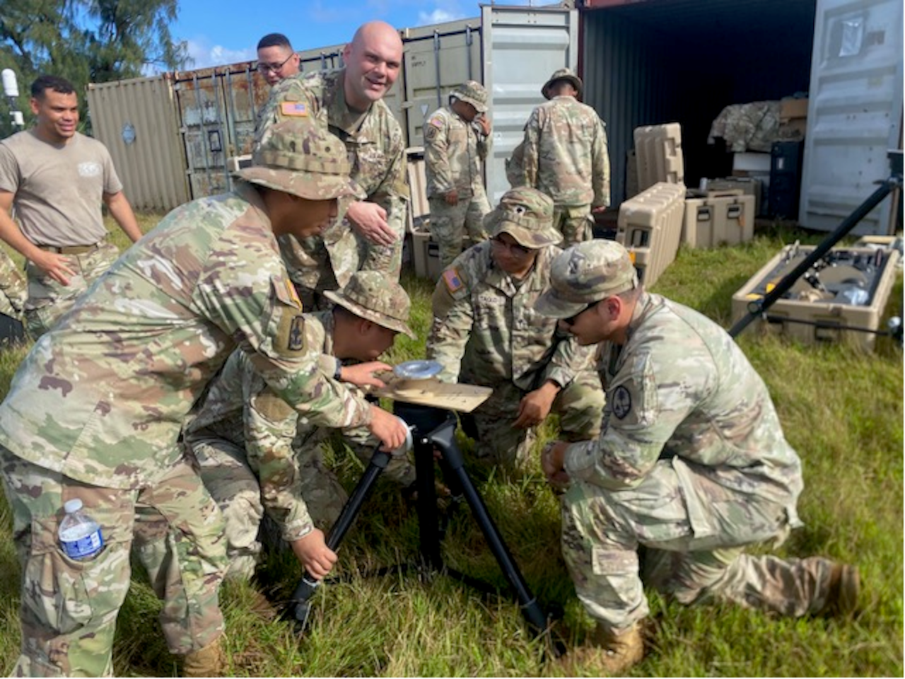 About six people in camouflage military uniforms kneel and stand in tall grass under a blue sky, looking at and working on a small drone that sits on a tripod. In the background, people in similar attire stand near large military storage containers.