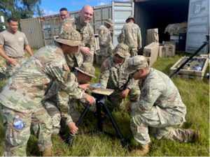 About six people in camouflage military uniforms kneel and stand in tall grass under a blue sky, looking at and working on a small drone that sits on a tripod. In the background, people in similar attire stand near large military storage containers.