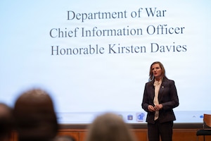 A woman in business attire stands on a stage speaking to an audience that is mostly off-screen. Behind her is a slide projected on the wall that reads, “Department of War Chief Information Officer Honorable Kirsten Davies.”