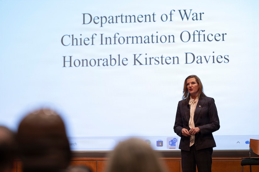 A woman in business attire stands on a stage speaking to an audience that is mostly off-screen. Behind her is a slide projected on the wall that reads, “Department of War Chief Information Officer Honorable Kirsten Davies.”